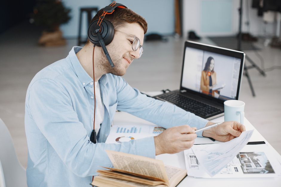 Homem trabalhando remotamente com fones de ouvido, revisando documentos e fazendo videoconferência no laptop.