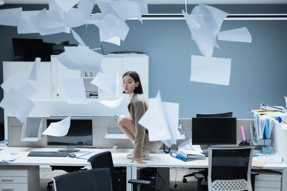 Mulher empoleirada em uma mesa em um escritório caótico com papéis voando, representando o estresse no trabalho.