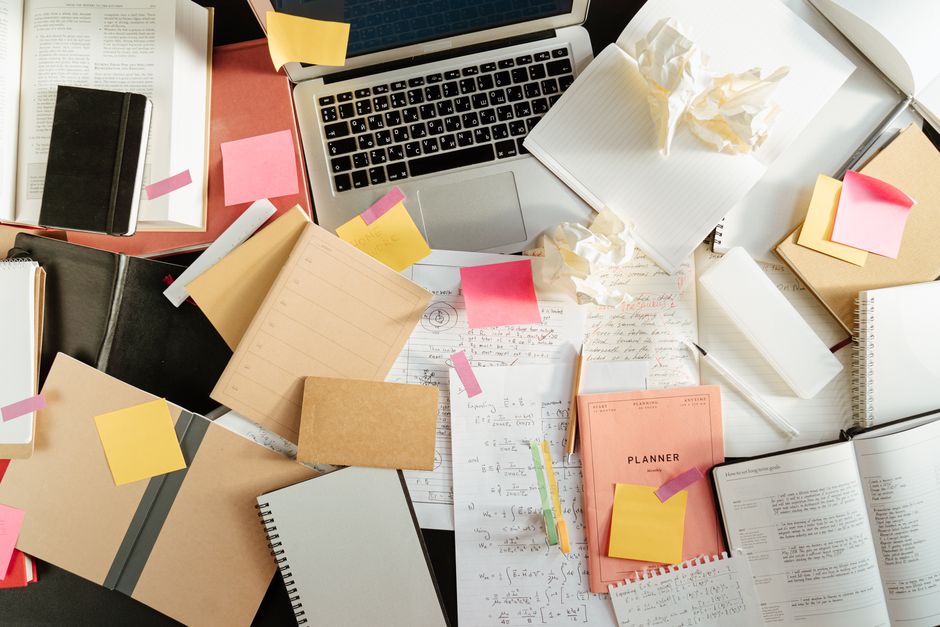 Foto aérea de uma mesa de estudo bagunçada com laptop, cadernos e notas autoadesivas, mostrando o processo criativo.