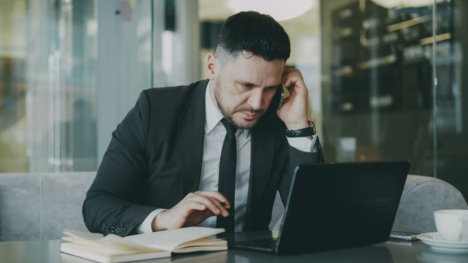 Empresário concentrado, de terno, realizando multitarefas no celular e no laptop em um escritório moderno.